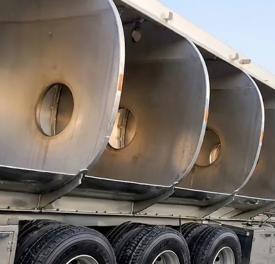 Underside view of a multi-compartment stainless steel tank trailer showing circular welded-baffle access ports between compartments